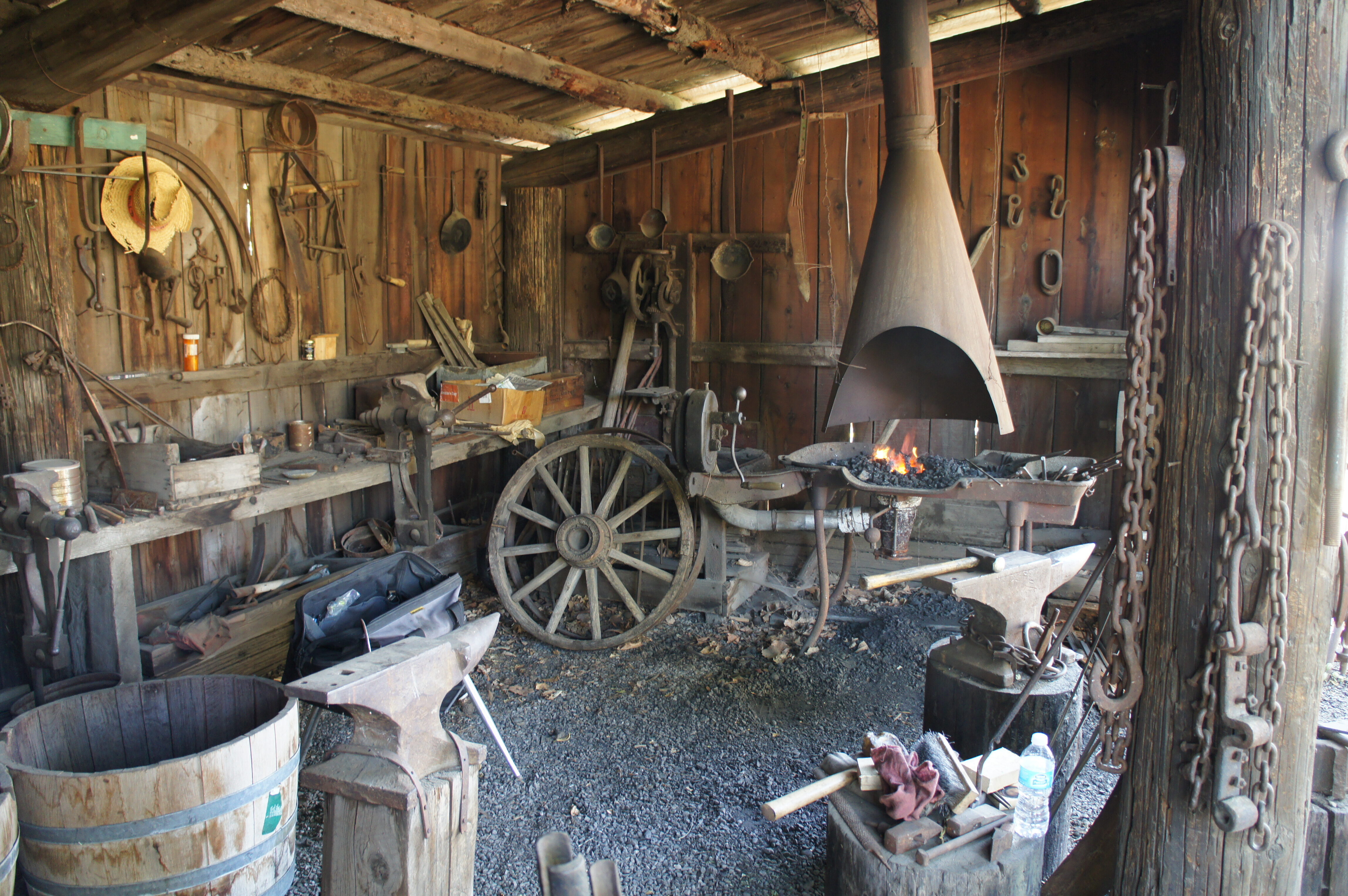The inside of a blacksmith's shop with a lit forge, wagon wheels, an anvil and tools.